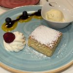 Un trozo cuadrado de tarta espolvoreada con azúcar glas en un plato azul, acompañado de una bola de helado de vainilla en un cuenco blanco, una quenelle de crema con una frambuesa encima y un chorrito de salsa de mango y frutos rojos.