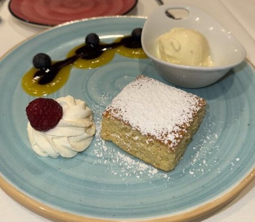 Un trozo cuadrado de tarta espolvoreada con azúcar glas en un plato azul, acompañado de una bola de helado de vainilla en un cuenco blanco, una quenelle de crema con una frambuesa encima y un chorrito de salsa de mango y frutos rojos.